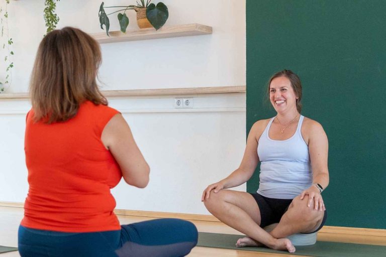 Lächelnde Yoga Schülerin sitzt im Schneidersitz einer anderen Schülerin gegenüber